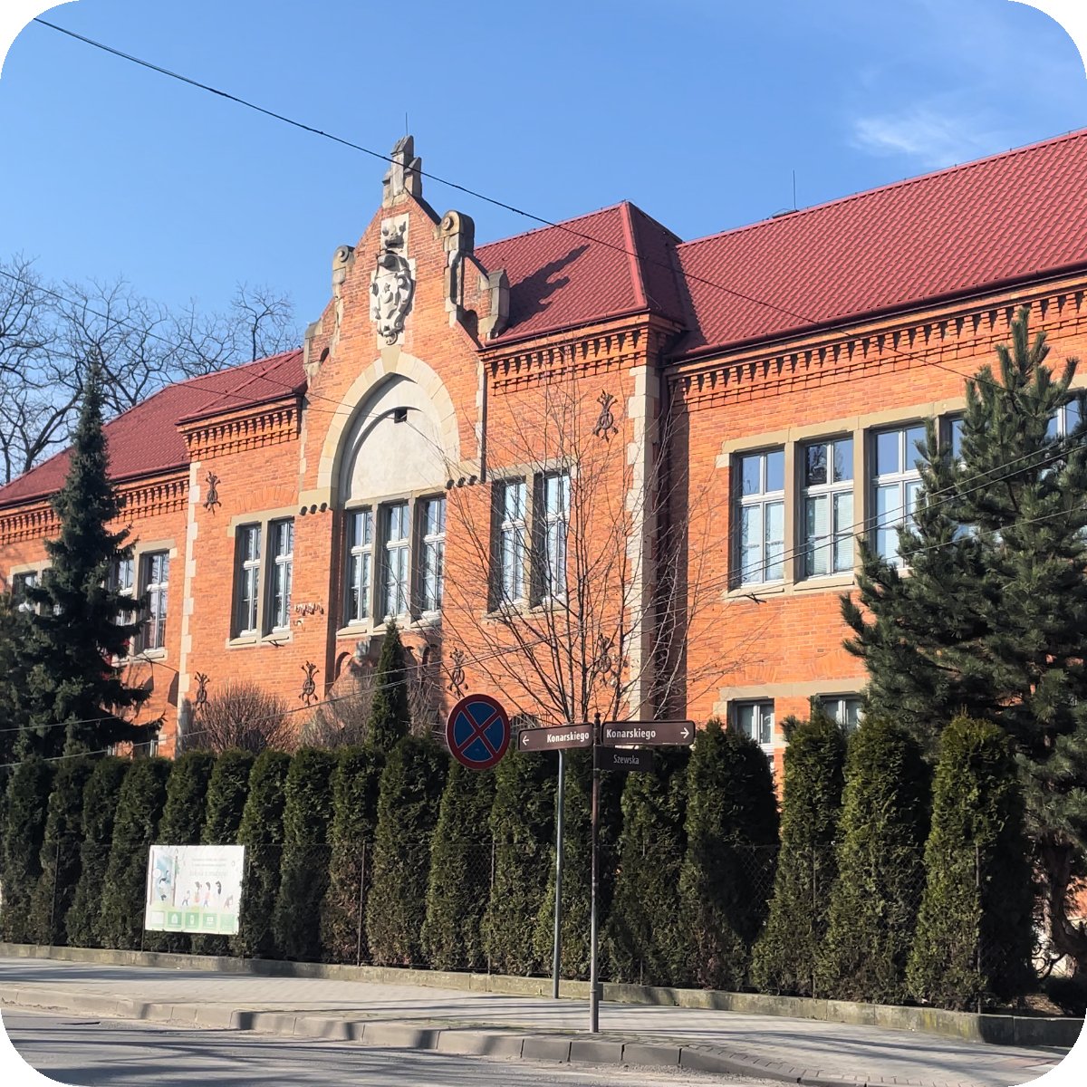Zdjęcie szkoły, pomarańczowy budynek zbudowany z cegieł lekko zasłonięty roślinnością, na pierwszym planie można dostrzec kawałek ulicy i chodnik